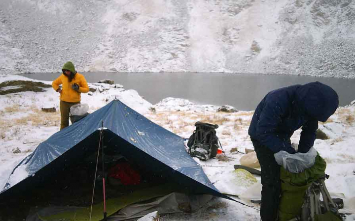 Two people stand near a tarp shelter that is resting beside a body of water. I light dusting of snow is covering the landscape.